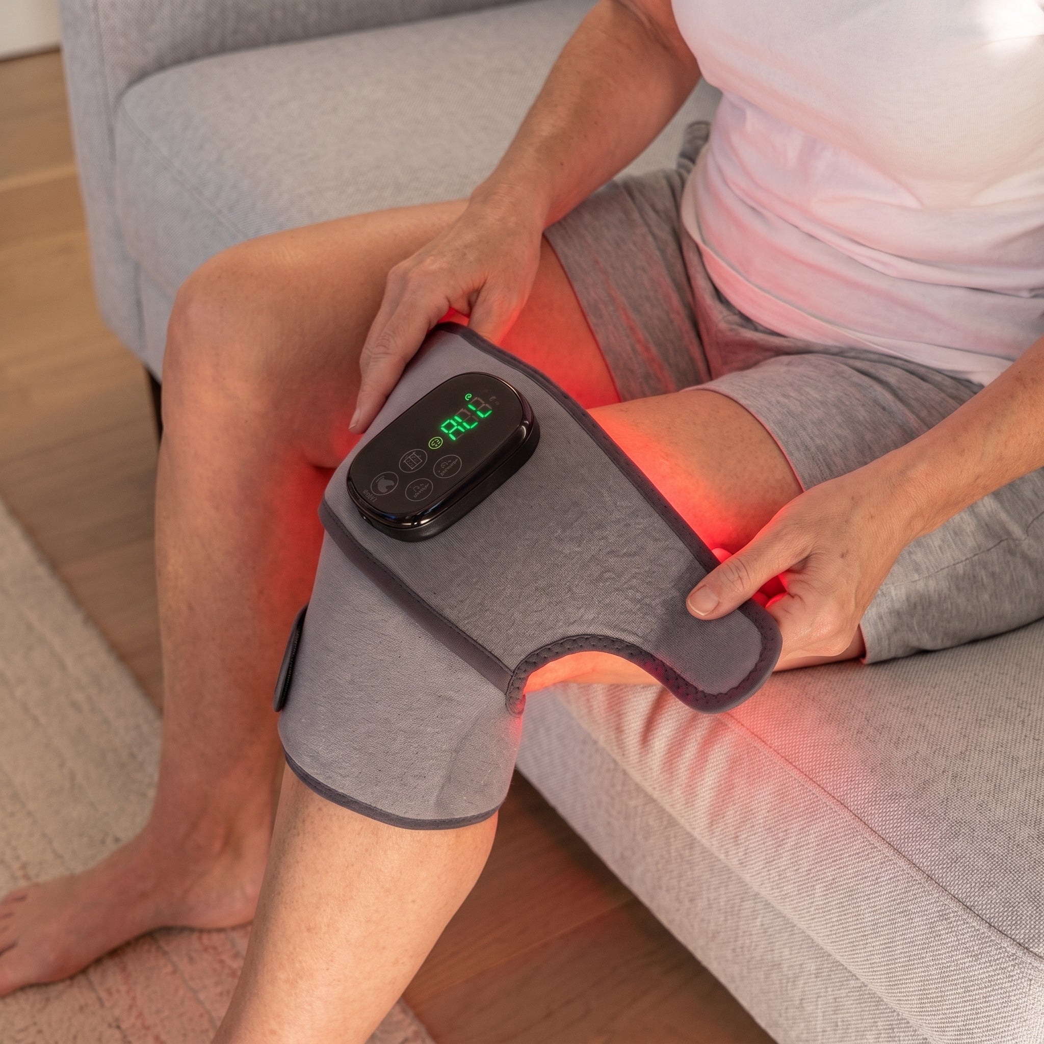 Person sitting on a couch wearing the LumoRelief Heat Therapy Brace on their knee, red light visible through the fabric, with the digital control display active